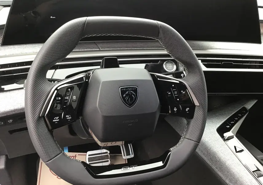 Vue rapprochée du volant cuir noir perforé du Peugeot 3008 1.2 Hybrid 145 GT+ avec tableau de bord numérique et bouton start/stop.