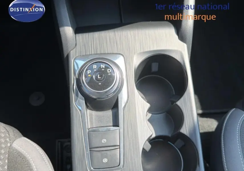 Vue rapprochée du sélecteur rotatif de boîte automatique et porte-gobelets sur la console centrale grise d’une Ford Focus SW bleu foncé.