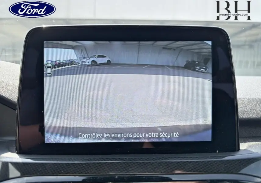 Écran tactile intérieur montrant la caméra de recul du Ford Kuga bleu, avec vue arrière sur parking.