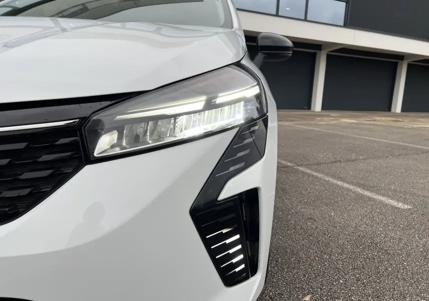 Gros plan sur le phare avant droit allumé d'une Renault Clio V blanche, soulignant ses feux LED modernes et le rétroviseur noir perle.