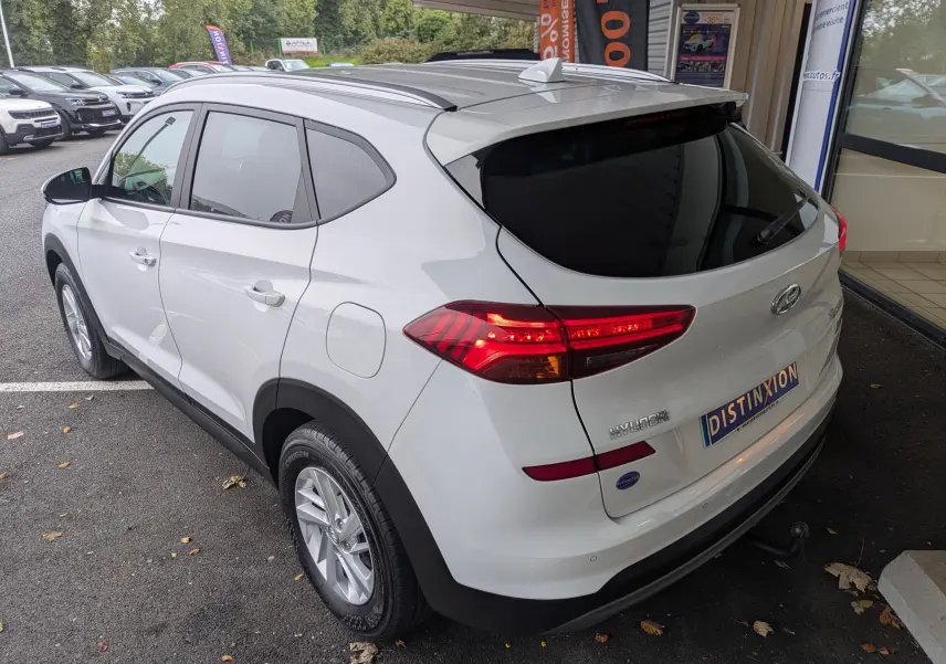 Vue 3/4 arrière droite d'un Hyundai Tucson blanc 2020 avec feux allumés et attelage visible en concession.