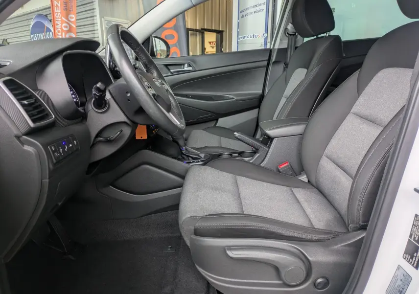 Vue intérieure côté conducteur du Hyundai Tucson 2020 blanc, sièges tissu gris et noir avec console centrale visible.