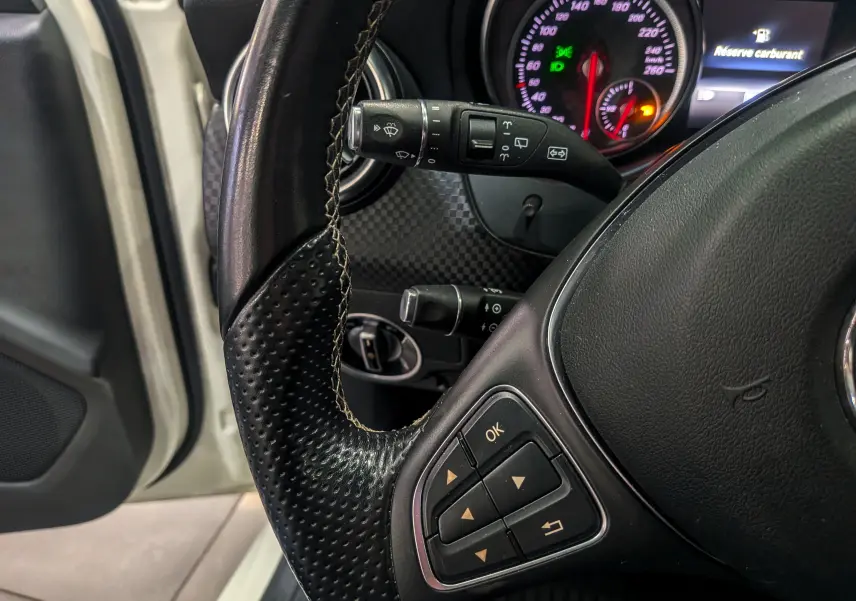 Gros plan sur le volant en cuir noir et le tableau de bord d'une Mercedes Classe A blanche, avec affichage digital et témoins allumés.