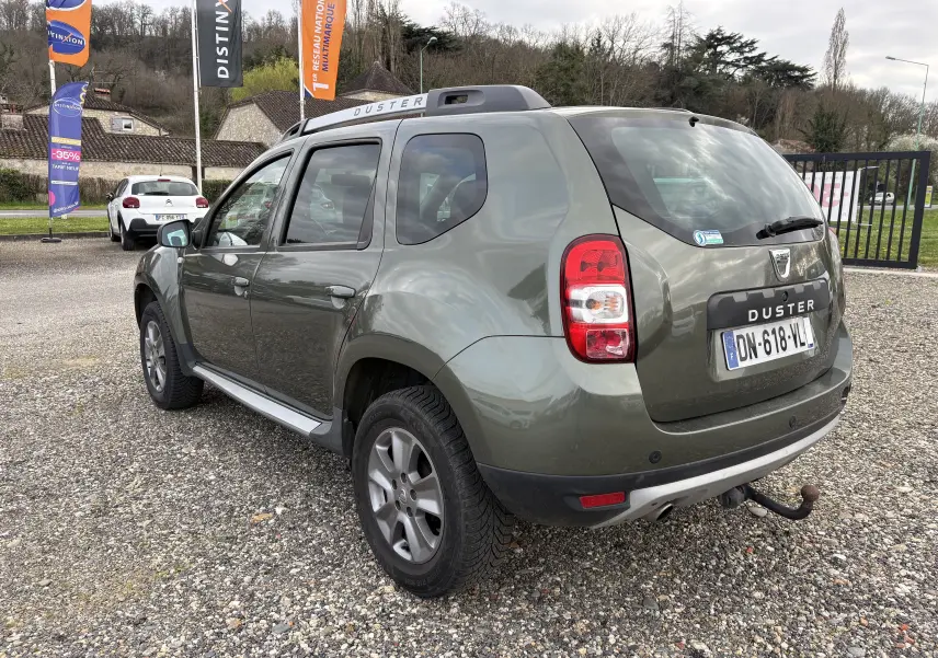 Vue 3/4 arrière droite d'un Dacia Duster vert foncé de 2014 avec attelage et barres de toit visibles.