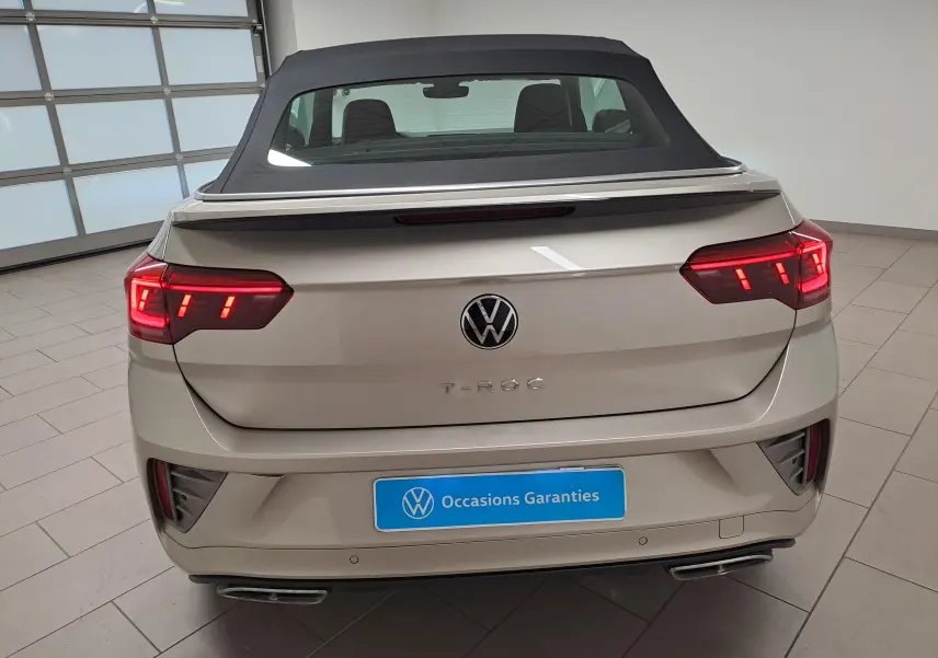 Vue arrière du Volkswagen T-Roc Cabriolet gris ivoire avec capote noire et feux arrière allumés dans un showroom.