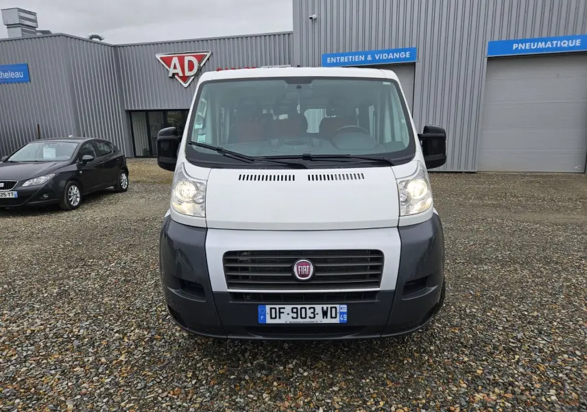 Vue de face d'un Fiat Ducato Combi blanc avec pare-chocs noir, immatriculé en France, stationné devant un garage.
