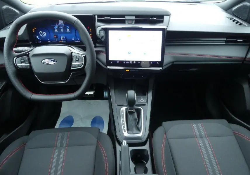 Intérieur Ford Puma 2025 vu de face, tableau de bord moderne avec écran tactile 12", volant et sièges noirs à surpiqûres rouges.