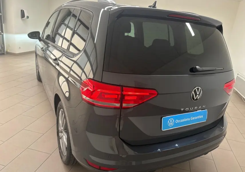 Vue 3/4 arrière droite d'un Volkswagen Touran gris dauphin 2025 avec feux arrière LED et hayon fermé dans un showroom.