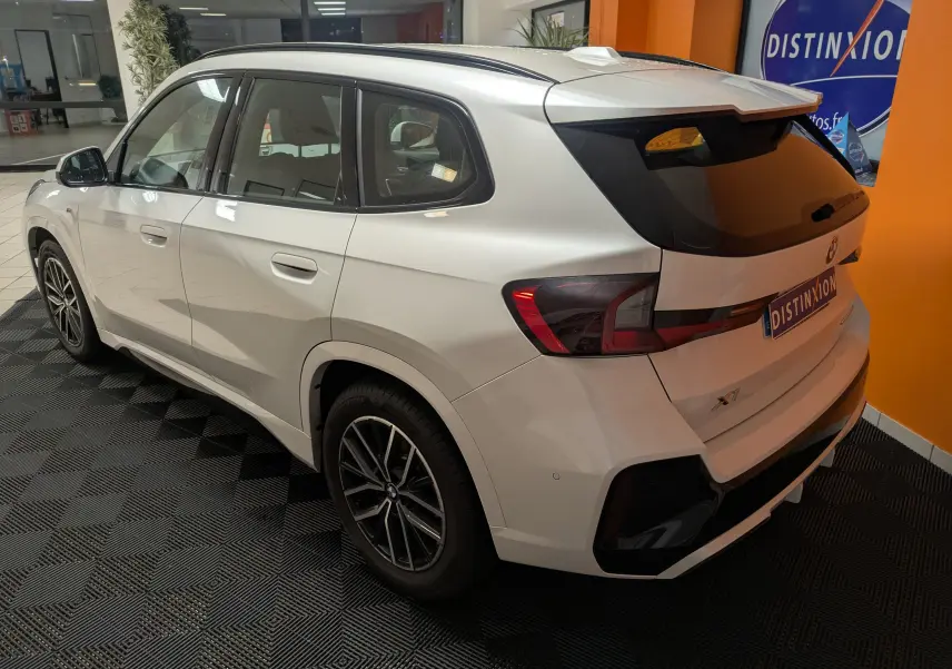 Vue 3/4 arrière droite d’un BMW X1 blanc 2025 avec jantes bicolores et vitres arrière teintées en intérieur showroom.