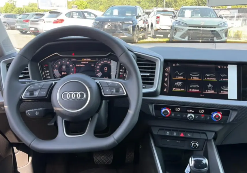 Vue intérieure centrée sur le volant multifonction Audi et l'écran tactile du tableau de bord, avec vue extérieure sur un parking.