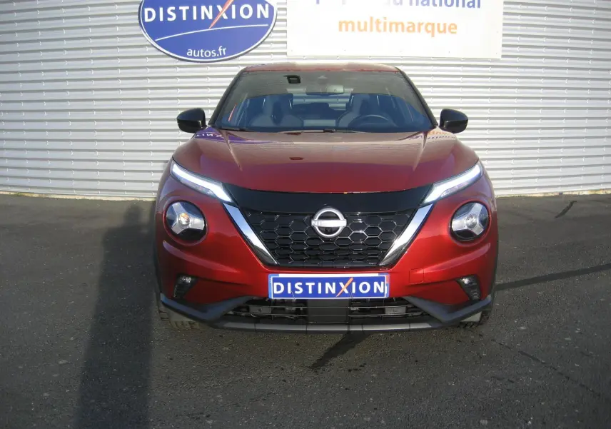 Vue frontale d'un Nissan Juke 1.6 Hybrid 143 Tekna rouge, avec calandre noire et phares LED distinctifs allumés.