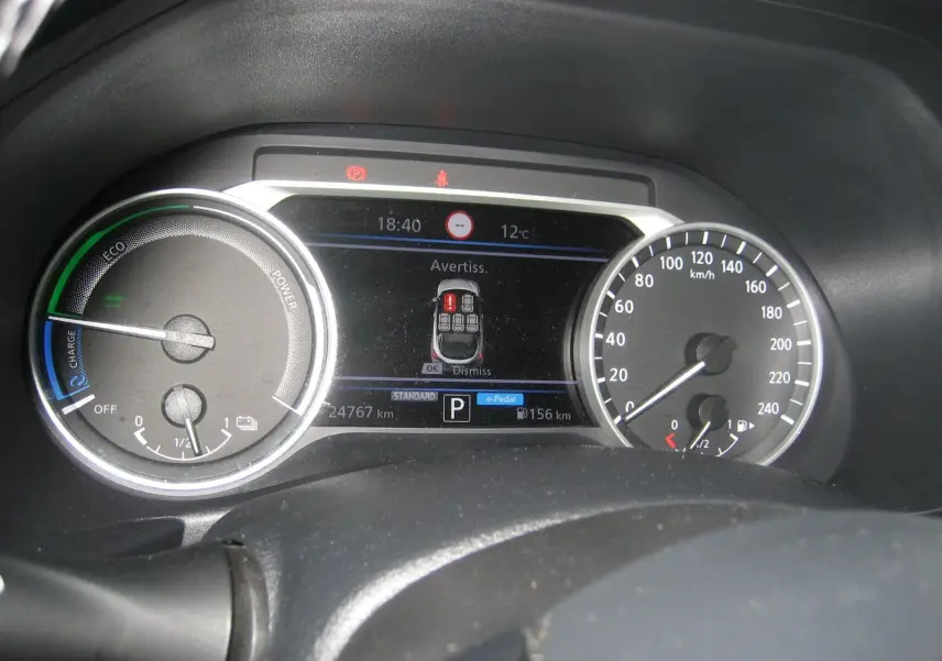 Vue rapprochée du tableau de bord numérique du Nissan Juke 1.6 Hybrid 143 Tekna, affichant l'alerte et la vitesse.