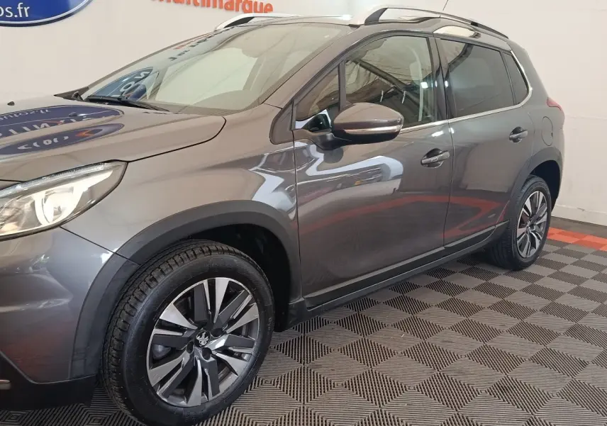 Vue 3/4 avant droit d’un Peugeot 2008 gris foncé avec jantes alliage et barres de toit chromées en intérieur showroom.