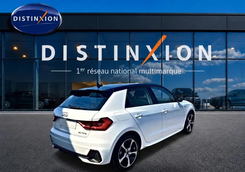 Audi A1 Sportback blanc et noir en 3/4 arrière droit, avec jantes alu et becquet arrière visible devant un showroom Distinxion.