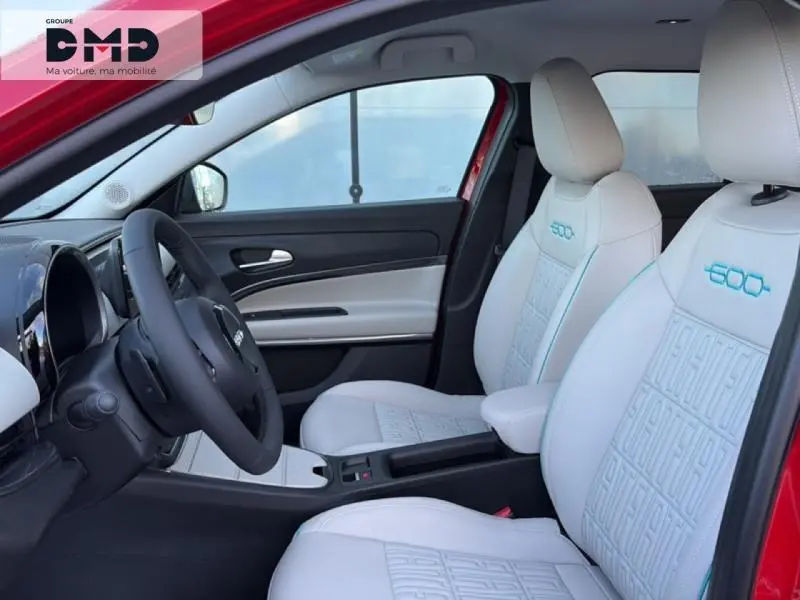 Vue intérieure côté conducteur d'une FIAT 600 rouge, sièges blancs avec logo 600 brodé bleu et tableau de bord noir.