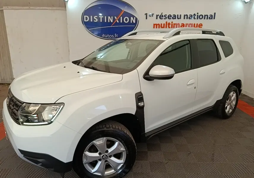 SUV Dacia Duster blanc en 3/4 avant droit, avec barres de toit noires et jantes alliage visibles.