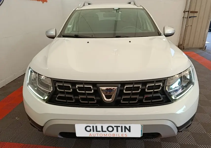 Vue avant d'un Dacia Duster blanc avec calandre noire et phares LED allumés en intérieur showroom.