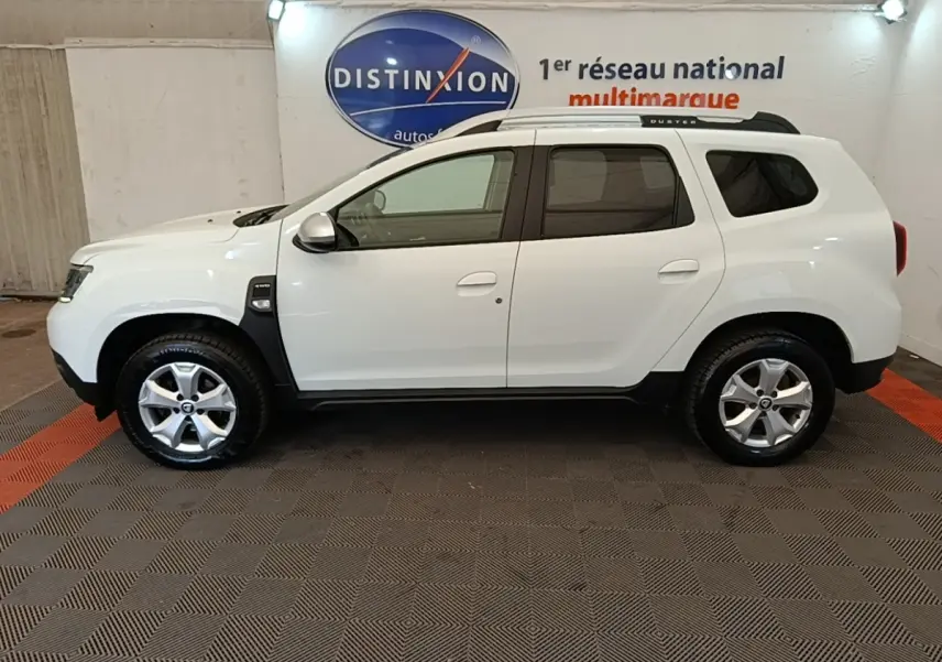 Vue de profil droit du Dacia Duster blanc 2021 avec barres de toit noires et jantes alliage en intérieur showroom.