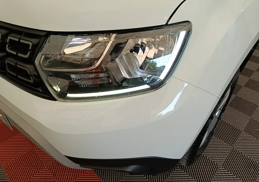 Gros plan sur le phare avant droit du Dacia Duster blanc, soulignant la calandre noire et le pare-chocs gris.