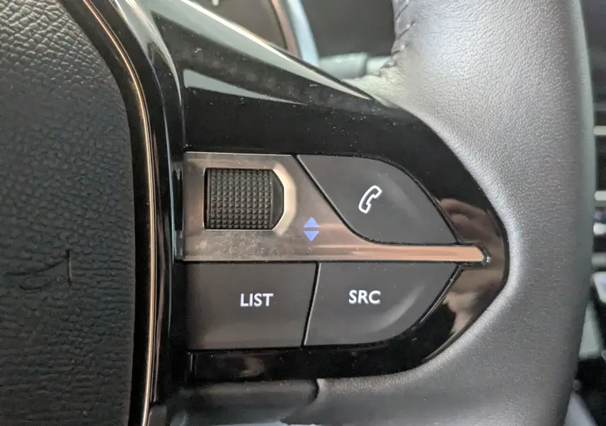 Gros plan sur les commandes gauche du volant noir du Peugeot 2008 Hybrid 145 e-DCS6 Allure, boutons SRC et LIST visibles.