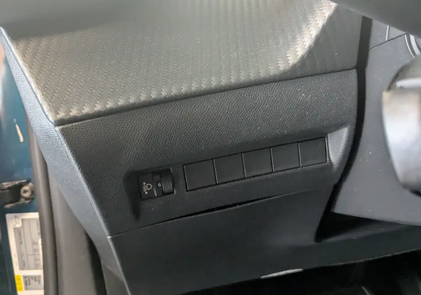 Détail intérieur côté gauche du tableau de bord noir texturé avec commande de réglage des phares sur Peugeot 2008 Bleu Obsession.