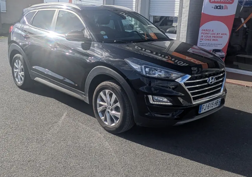 Hyundai Tucson noir vu en 3/4 avant droit, avec calandre chromée et jantes alliage, stationné devant un bâtiment.