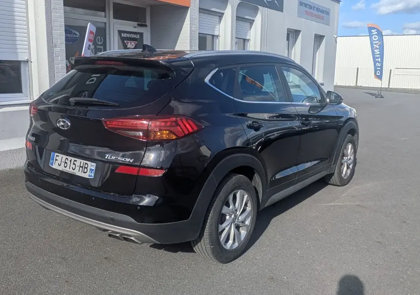 Vue 3/4 arrière droite d'un Hyundai Tucson noir garé devant un bâtiment commercial avec plaques d'immatriculation françaises.