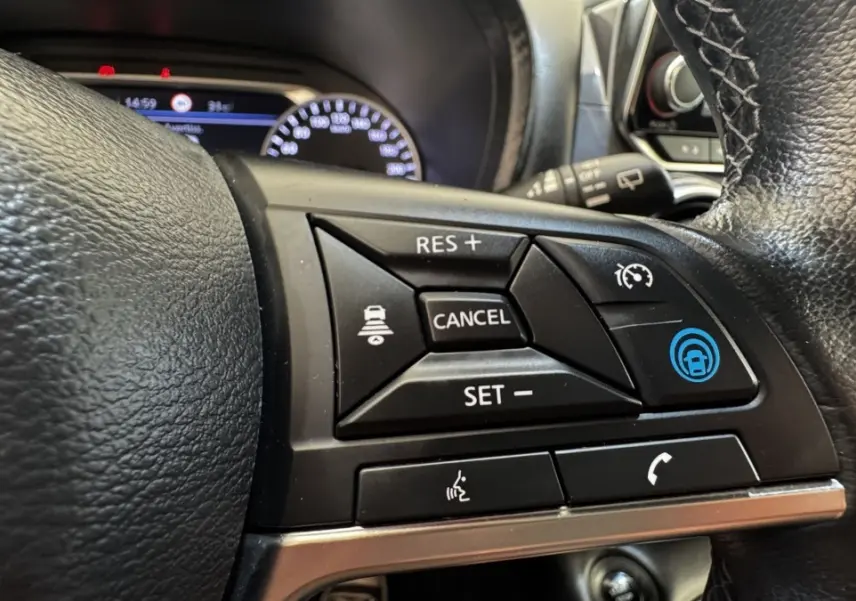 Gros plan sur les commandes du volant noir du Nissan Juke Hybrid 143 Business+ 2023, affichage du combiné en arrière-plan.