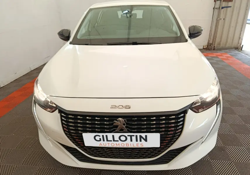 Vue avant d'une Peugeot 208 blanche 2023 avec calandre noire et phares allumés dans un garage.