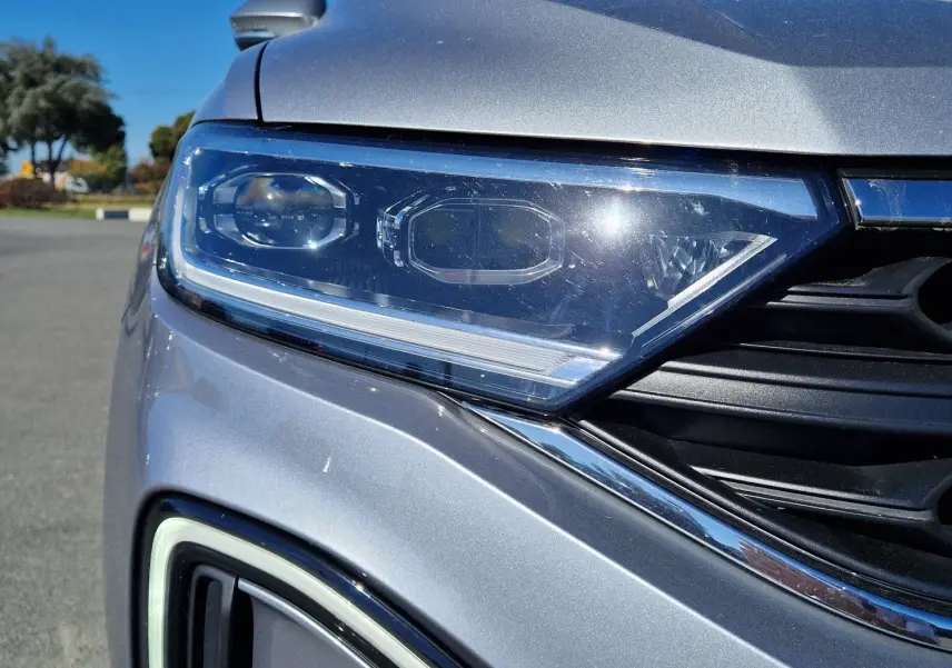 Gros plan sur le phare avant gauche et la calandre d'un Volkswagen T-Roc gris pyrite sous un ciel bleu.