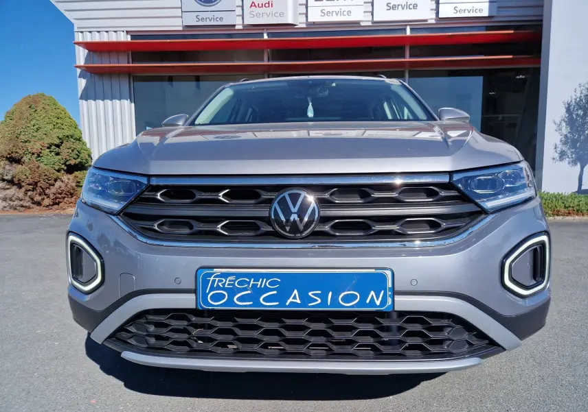 Vue frontale d'un Volkswagen T-Roc gris pyrite 2023 avec calandre noire et feux LED distinctifs allumés.