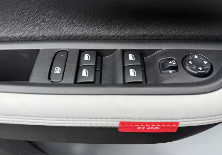 Détail des commandes de lève-vitres et rétroviseurs sur la porte intérieure d'une Citroën C3 hybride gris Mercury avec surpiqûres blanches et étiquette rouge "be cool".