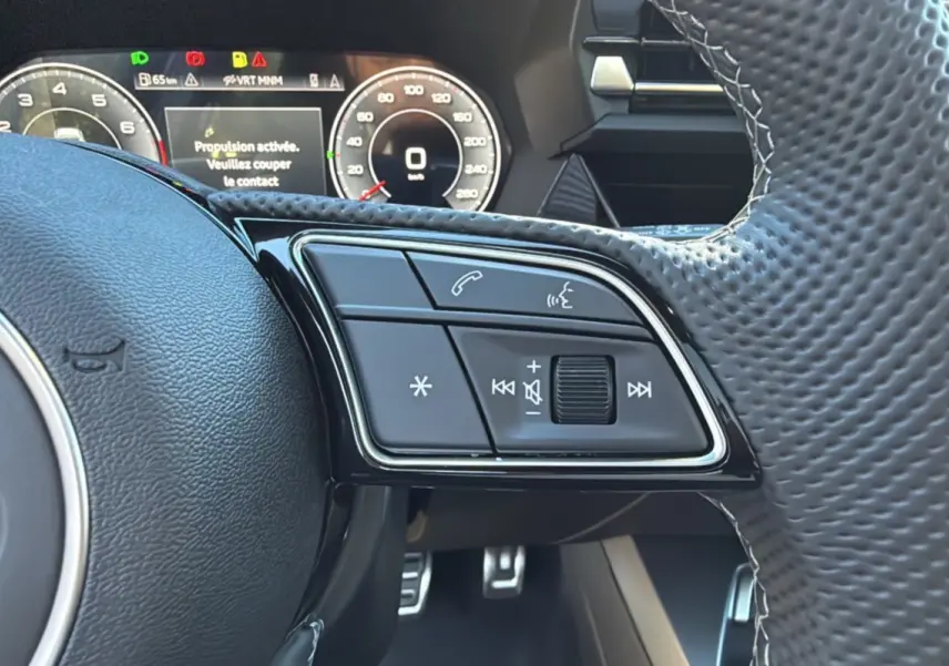 Gros plan sur les commandes au volant à méplat de l'Audi A3 TFSI 150 Hybrid S-Line 2025 avec tableau de bord numérique.