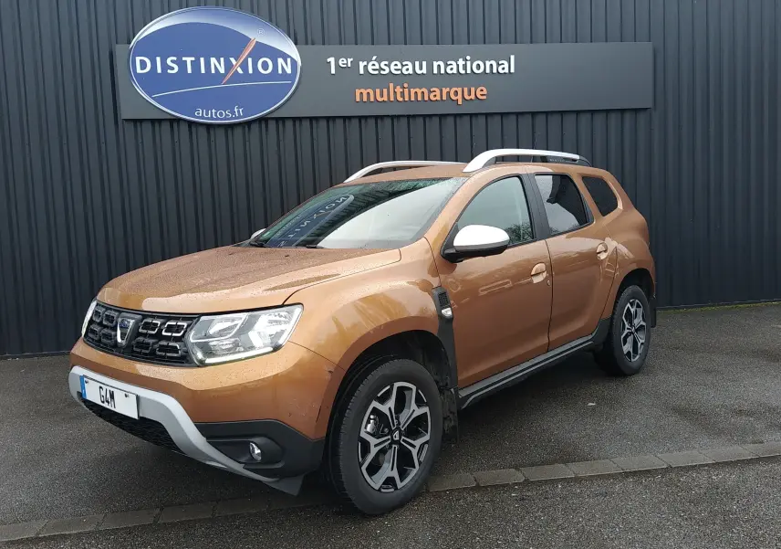 Dacia Duster orange Atacama en 3/4 avant droit, avec jantes alliage et barres de toit blanches visibles.