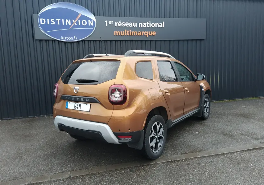Vue 3/4 arrière droite du Dacia Duster orange Atacama 2021 avec jantes alliage et barres de toit visibles.