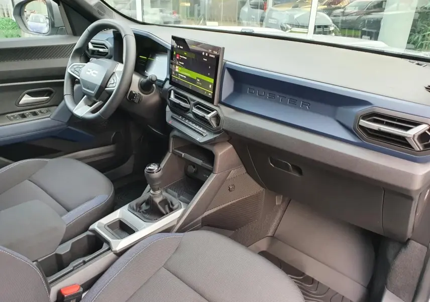 Vue intérieure côté conducteur du Dacia Duster 2025, tableau de bord bleu, écran tactile et levier de vitesses manuel visible.