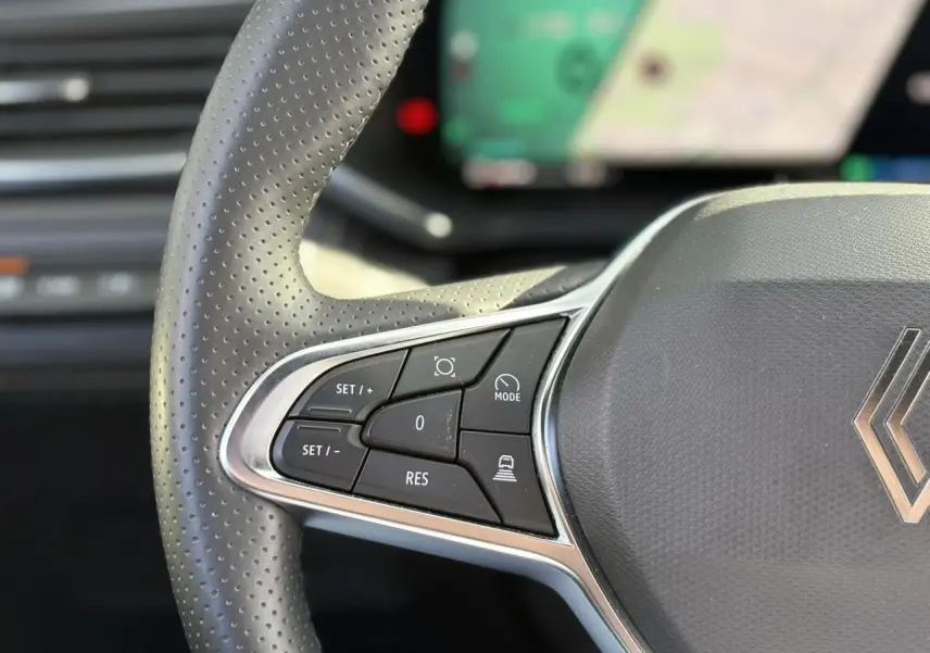 Gros plan sur les commandes à gauche du volant perforé gris du Renault Symbioz E-Tech Full Hybrid 2025 avec écran tactile flou en arrière-plan.