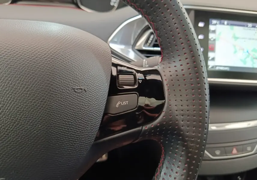 Gros plan sur le volant cuir noir perforé avec surpiqûres rouges de la Peugeot 308, écran tactile de navigation en arrière-plan.