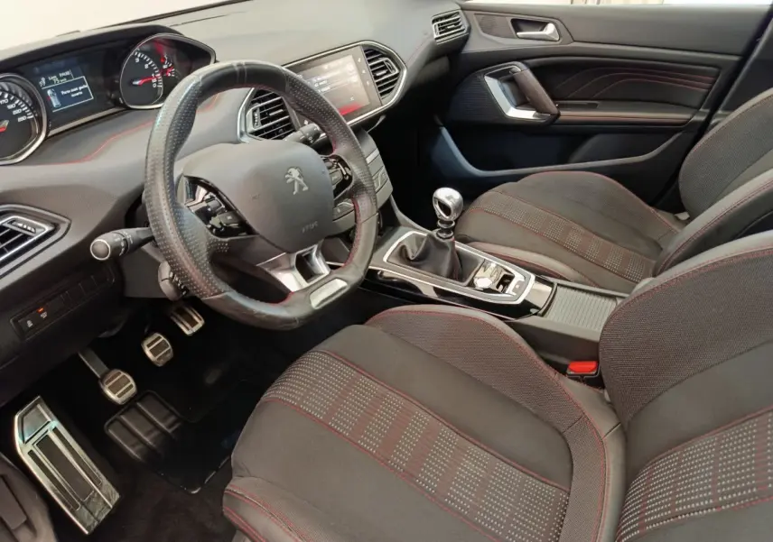 Intérieur Peugeot 308 GT Line 2016 vu côté conducteur, volant cuir, sièges tissu noirs avec surpiqûres rouges.