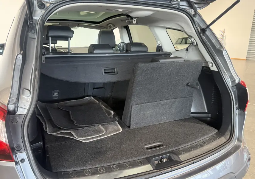 Coffre ouvert du Nissan Qashqai+2 gris 2014, vue arrière montrant la banquette arrière relevée et tapis de sol empilés.