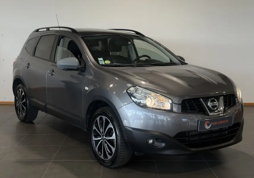 Nissan Qashqai+2 gris 2014 en 3/4 avant droit, avec jantes noires et phares allumés dans un intérieur sobre.