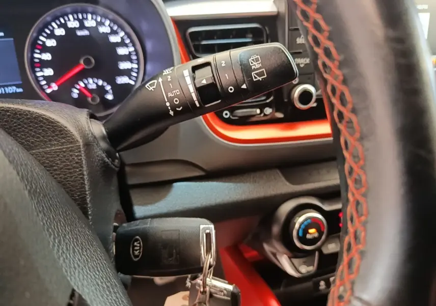 Gros plan sur le volant cuir noir avec surpiqûres rouges et commandes, tableau de bord et clés KIA visibles.