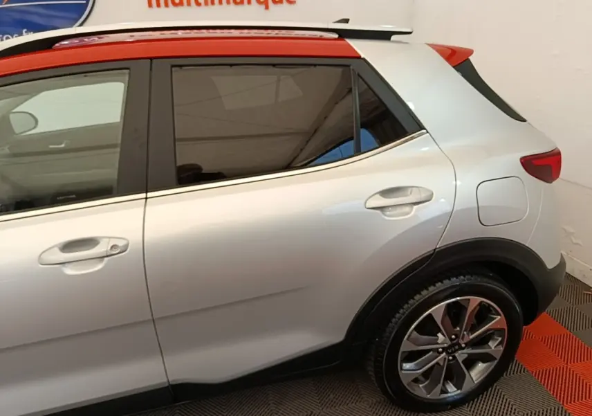 Vue latérale droite d'un KIA Stonic gris clair avec toit rouge et jantes alliage, en intérieur showroom.