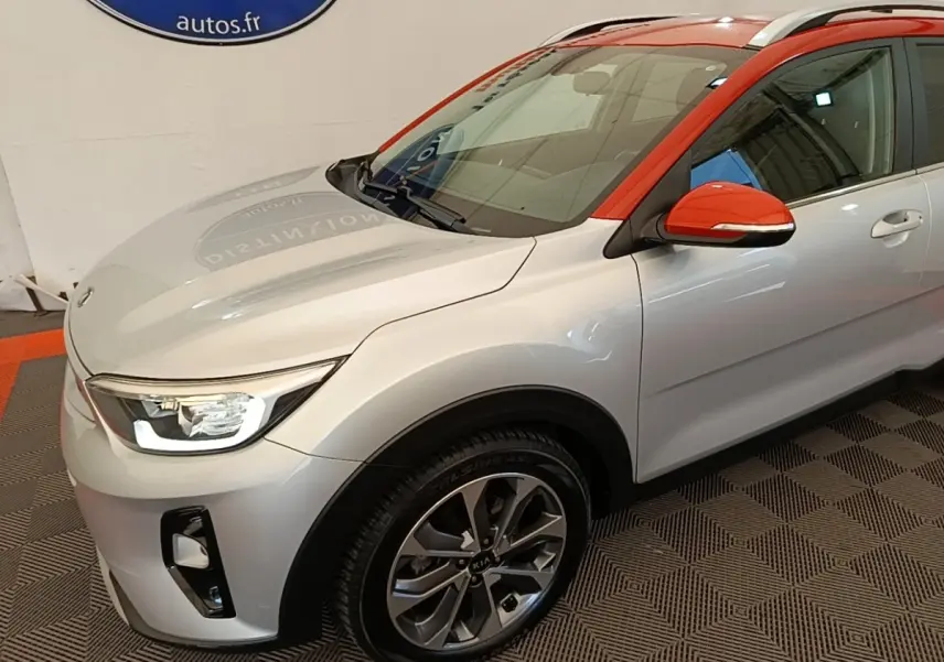 Vue 3/4 avant droit d’un KIA Stonic gris clair avec toit et rétroviseur orange vif en intérieur showroom.