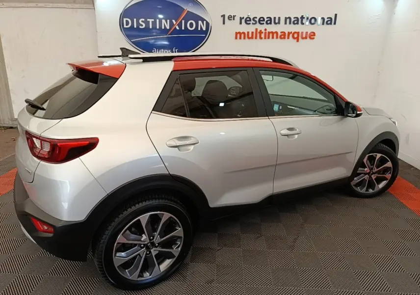 Vue 3/4 arrière droite d’un KIA Stonic gris clair avec toit rouge et jantes alliage, en intérieur showroom.