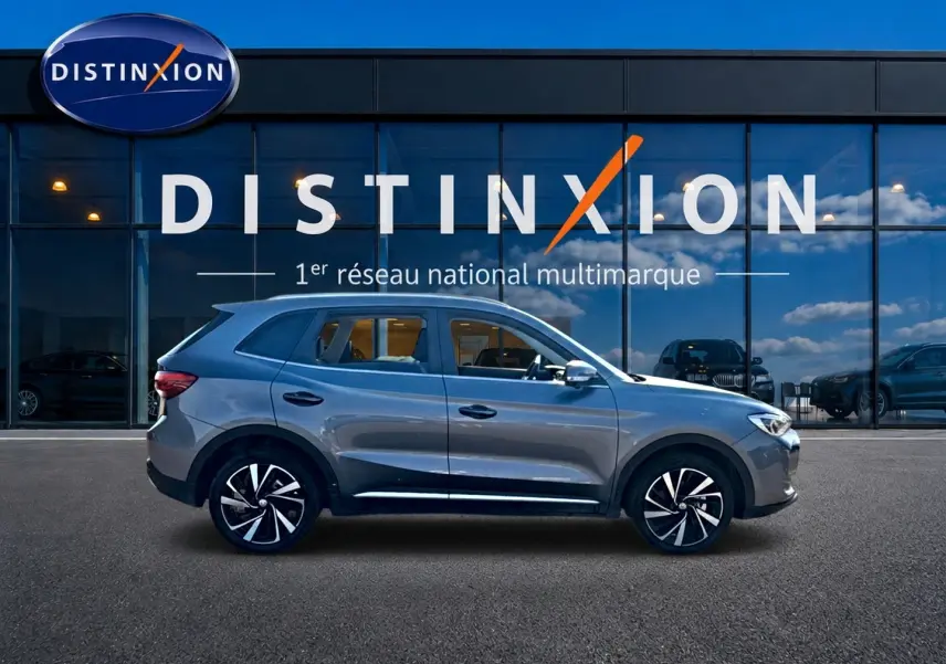 Vue de profil côté gauche du MG Motor ZS gris 1.5 hybrid 197 luxury devant une vitrine Distinxion.