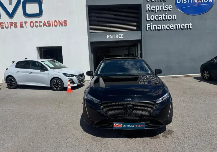 Vue de face d'une Peugeot 308 Hybrid 136 e-DCS6 Allure noire, stationnée devant un garage avec enseigne visible.