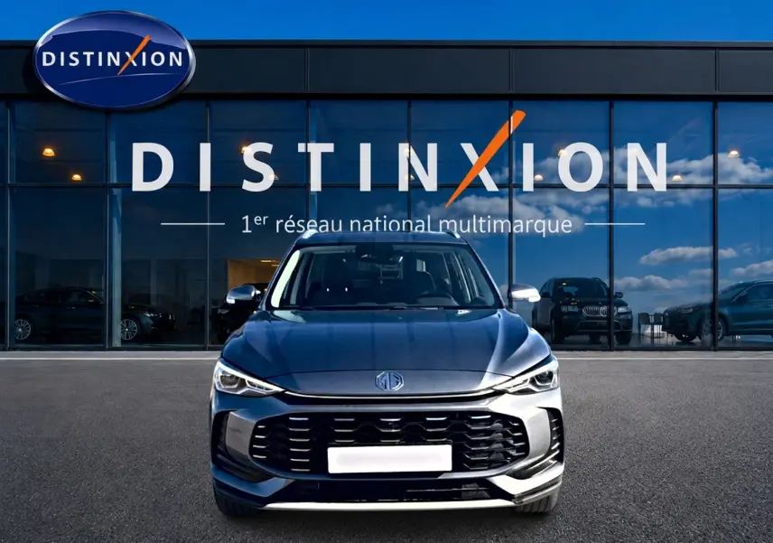 Vue frontale d'un MG MOTOR ZS 1.5 hybrid gris 2026 avec calandre noire et phares LED devant un showroom Distinxion.