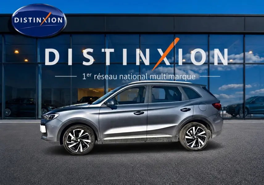 Vue de profil côté gauche d’un MG ZS gris 2026 hybride avec jantes alliage noires et blanches, devant une vitrine Distinxion.