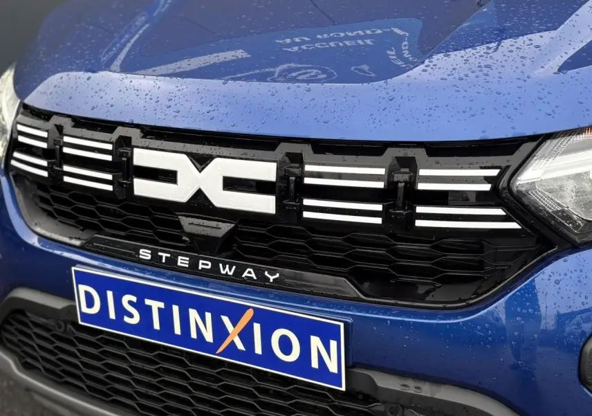 Gros plan sur la calandre avant noire et le logo Extrême blanc du Dacia Sandero Stepway Bleu Iron avec pluie visible.
